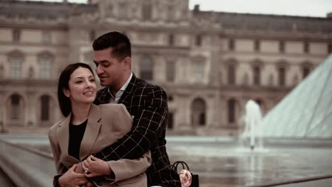Elegant-couple-sitting-and-swinging-while-hugging-romantic-at-the-Museum-du-Louvre-Pyramid-and-fountain-surrounded-by-the-baroque-royal-residence-buildings-in-Paris-France---modern-lifestyle-and-suits