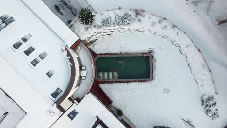 Aerial-drone-shot-of-luxurious-fancy-hotel-and-spa-resort-in-middle-of-mountains,-covered-in-winter-snow-with-amazing-swimming-pool-with-steaming-water,-people-relaxing-after-skiing