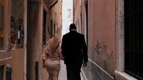 A-classy-mixed-race-couple-walking-through-the-narrow-streets-of-Venice-Italy-between-the-typical-old-houses-to-explore-the-city-while-traveling---view-from-the-back