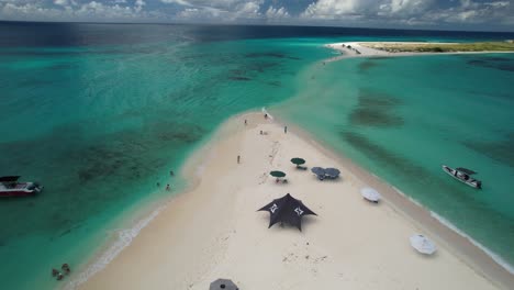 Aerial-zoom-in-cayo-de-agua-island,-people-enjoy-day-beach,-white-sand,-boats-and-beach-umbrellas