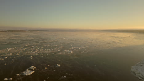 Backwards-drone-view-going-towards-the-coast-of-the-Canadian-town-of-Beaumont,-Quebec,-from-the-St-Lawrence-river-Seaway,-on-a-very-cold-Winter-morning