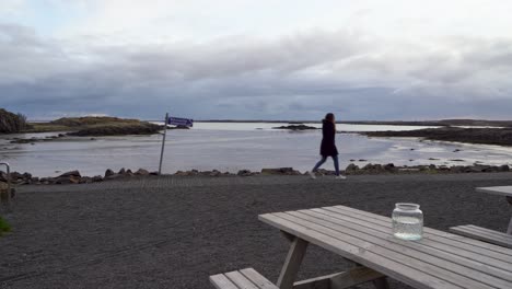 Frau-Geht-An-Einer-Ruhigen-Isländischen-Küste-Entlang,-Bewölkter-Himmel,-Reflektierendes-Wasser,-Mit-Picknicktischen-Im-Vordergrund