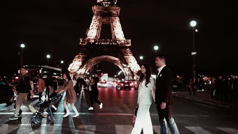 Elegant-dark-haired-couple-in-black-and-white-suit-walking-over-the-street-at-Pont-d´lena-in-front-of-the-famous-Eiffel-Tower-at-night-while-all-the-people-and-traffic-lights-around-take-some-photos