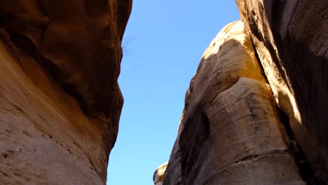 Looking-up-at-steep,-craggy-red-rock-formations-at-sunny-blue-sky-deep-within-valley-in-the-desert-wilderness-of-Wadi-Ghuweir-in-Jordan,-Middle-East