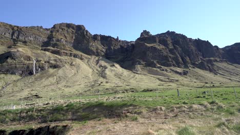 üppig-Grüne-Isländische-Landschaft-Mit-Schroffen-Klippen-Und-Einem-Kleinen-Wasserfall-Unter-Klarem-Blauen-Himmel