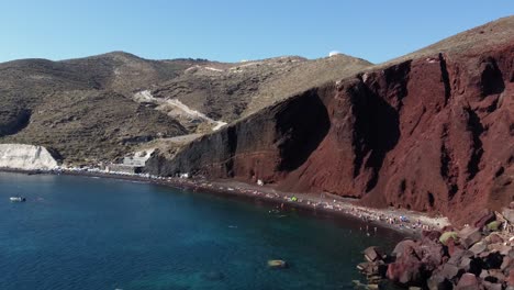 Aerial-Orbit-Red-Beach-full-of-Tourists-and-Steep-Cliff,-Santorini,-Greece