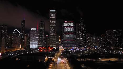 Vista-Aérea-Sobre-El-Parque-Del-Milenio,-Hacia-Los-Rascacielos-Iluminados-Por-La-Noche-En-El-Día-De-Vestir-Rojo-En-Chicago