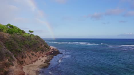 Drohnenaufnahmen-Entlang-Der-Von-Klippen-Gesäumten-Küste-Der-Insel-Oahu-Mit-Einem-Regenbogen,-Der-Das-Strahlend-Blaue-Wasser-Des-Pazifischen-Ozeans-Berührt