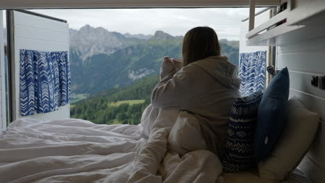 Beautiful-woman-having-cup-of-coffee-on-early-morning-in-bed-on-camper-van,-Dolomites-in-background