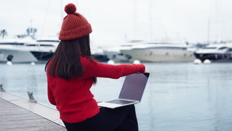 Eine-Asiatische-Frau-Sitzt-Am-Wasser-Im-Hafen-Von-Valencia-Und-öffnet-Ihre-Arbeit-Auf-Einem-Laptop,-Mit-Einer-Wunderschönen-Kulisse-Aus-Yachten,-Und-Trägt-Eine-Rote-Beanie,-Die-Ihr-Rotes-Rollkragenshirt-Ergänzt