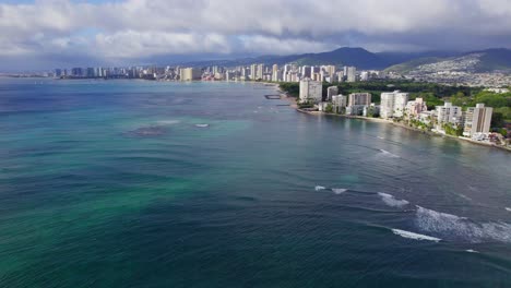 Drone-footage-above-the-aqua-blue-water-of-the-Pacific-Ocean-along-the-coast-of-the-island-of-Oahu-,Hawaii-Honolulu-cityscape-lines-the-coast-with-volcanic-formations-on-the-horizon
