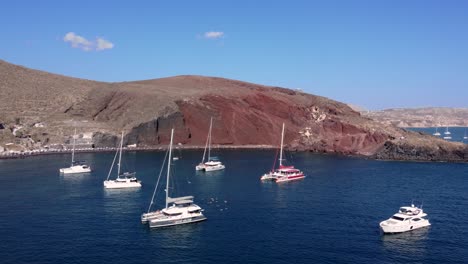Orbital-Shot-of-Red-Beach-Bay-with-Catamarans-and-Tourists-Swimming,-Santorini,-Greece