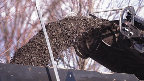 Zeitlupenaufnahme-Von-Zerkleinertem-Straßenasphalt,-Der-Aus-Einer-Maschine-Auf-Die-Ladefläche-Eines-Baufahrzeugs-Befördert-Wird,-Während-Der-Verkehr-Auf-Der-Gegenfahrbahn-Fährt