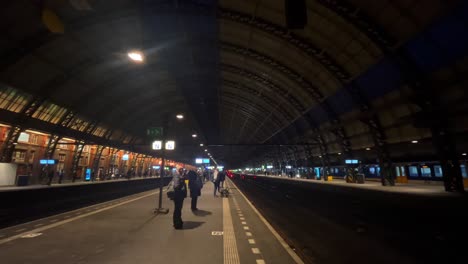 Gente-Esperando-En-El-Andén-Oscuro-De-La-Estación-Central-De-Trenes-De-Amsterdam-Por-La-Noche