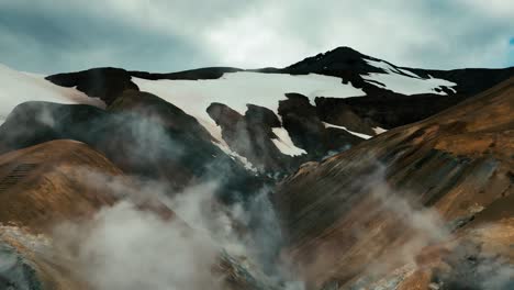 Aerial-drone-flying-toward-a-series-of-steaming-geysers-and-hot-springs-amid-snow-capped-mountains-and-hills