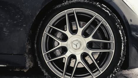 Close-up-of-parked-black-car-metallic-tire-rim,-snowy-ground-and-bright-sky