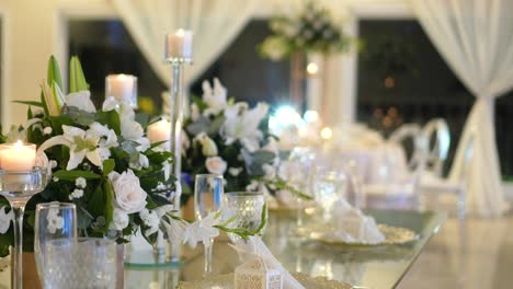 Glass-table-decorated-with-centerpieces-with-natural-foliage-and-white-roses,-candlesticks-and-glassware-for-wedding-reception