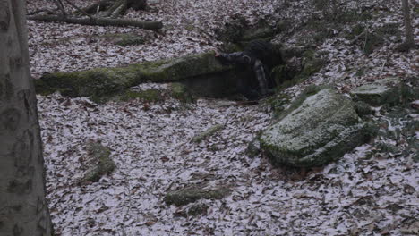 Ein-Wanderer-Klettert-An-Einem-Wintertag-Mit-Einer-Leichten-Schneeschicht-Aus-Einer-Dunklen-Höhle,-Die-Zwischen-Den-Felsbrocken-Versteckt-Ist,-Auf-Den-Mit-Blättern-Bedeckten-Waldboden