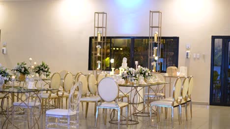 Sala-De-Reuniones-Decorada-Para-Una-Boda,-Con-Mesas-De-Cristal-Con-Centros-De-Follaje-Y-Elegantes-Sillas-Doradas.