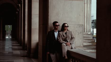 Pareja-Elegante-Con-Gafas-De-Sol-Negras-Apoyadas-Y-Sentadas-En-La-Pared-Entre-El-Arco-Disfrutando-De-La-Vista-Del-Lugar-Vacío-Alrededor-Del-Museo-Del-Louvre-Con-Sus-Edificios-Borque-En-París,-Francia