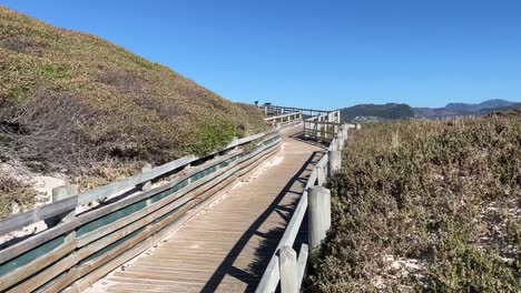 A-wooden-boardwalk-leading-to-Boulders-Beach-near-Simons-Town,-South-Africa,-pier-leads-through-brush-covered-sand-dunes