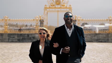 Classy-mixed-race-couple-with-sunglasses-walking-over-the-empty-Cour-d'Honneur---honor-courtyard-of-the-famous-castle-Versailles-in-Paris-France-with-the-golden-honour-gate-in-the-background-cinematic