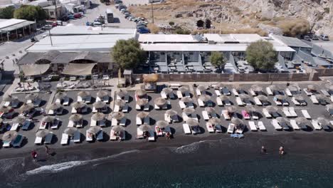 Aerial-Sliding-Shot-of-Perissa-Beach-full-of-Tiki-Huts-and-Tourists,-Santorini