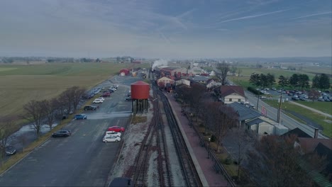 An-Aerial-View-of-a-Train-Station,-with-a-Steam-Passenger-Train-Approaching-in-the-Distance,-on-a-Partially-Sunny-Day