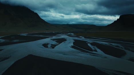 Drone-aerial-of-a-twisting-snakelike-river-in-the-cool-dark-of-the-ominous-clouds-and-mountain-range
