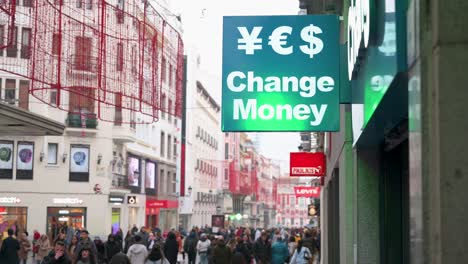 On-a-busy-and-crowded-retail-commercial-street,-a-currency-exchange-sign-shows-the-message-'Change-Money'-with-symbols-for-currencies-like-the-Japanese-yen,-US-dollar,-and-euro