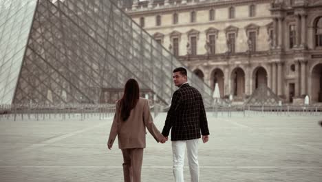 Elegantes-Paar,-Das-über-Den-Leeren-Platz-Des-Louvre-Museums-Geht-Und-Zurück-In-Die-Kamera-Blickt,-Umgeben-Von-Den-Barocken-Gebäuden-Der-Königlichen-Residenz-In-Paris,-Frankreich---Moderner-Lebensstil-Und-Passt-Zu-Verabredungen-Mit-Liebenden