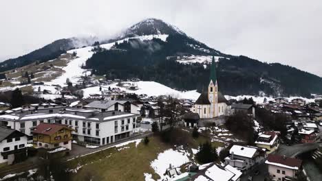 Snowy-mountain-peak-and-township-with-church-tower-of-Kirchberg,-aerial-drone-view