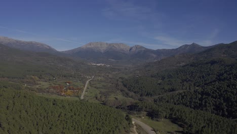 reverse-flight-in-a-valley-with-lush-pine-forests-and-in-the-background-large-mountains-with-a-road-with-cars-in-circulation-that-leads-to-a-mountain-pass-on-an-autumn-day-with-in-Avila-Spain