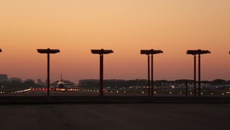Flugzeug-Startet-In-Der-Abenddämmerung-Mit-Landebahnbeleuchtung-Und-Stadtsilhouette-Im-Hintergrund,-Warmer-Himmel