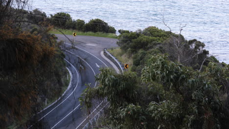 Aerial-view-of-a-car-driving-along-a-curved-road-surrounded-by-lush-foliage,-with-the-sea-nearby