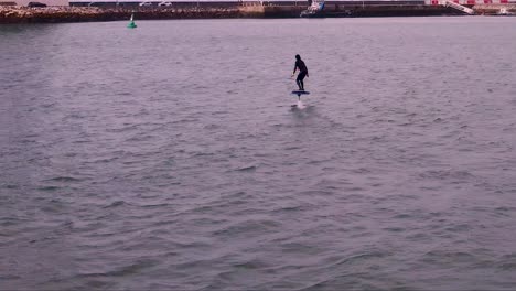 Luftbildverfolgung-Eines-Jungen-Mannes,-Der-Auf-Einem-Elektrischen-Hydrofoil-Surfbrett-Auf-Glasklarem-Blauem-Ozeanwasser-Reitet,-Mit-Einem-Steg-Hinter-Ihm