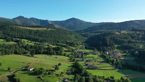 Vuelo-De-Drones-En-Un-Valle-Viendo-Un-Pueblo-Con-Sus-Casas-Blancas-Con-Techos-Rojos,-Campos-De-Cultivo-Y-Sus-Caminos-Con-Bosques-De-Pinos-Con-Montañas-Al-Fondo-En-Verano-Al-Atardecer-Icantabria-España