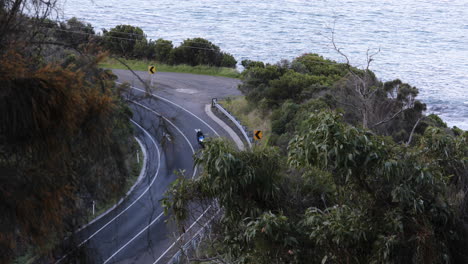 Aerial-view-of-a-motorcyclist-riding-along-a-curved-road-surrounded-by-lush-foliage,-with-the-sea-nearby