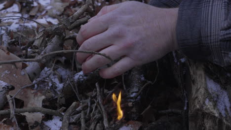 a-man-uses-his-hands-to-shield-the-tiny-flames-from-the-wind-as-he-begins-to-build-a-campfire-in-the-winter-with-leaves-and-snow-on-the-ground