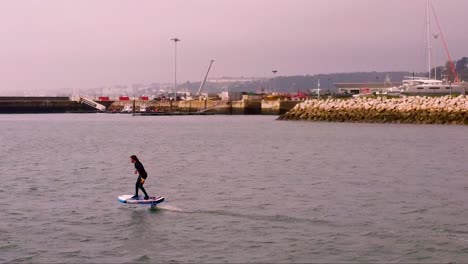 Aerial-view-of-a-young-man-riding-on-an-electric-hydrofoil-surfboard-on-glassy-blue-ocean-water-with-dock-behind-him