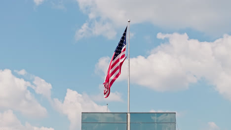 Ondeando-La-Bandera-Americana-En-El-Asta-De-La-Bandera-Contra-El-Cielo-Azul-Y-Las-Nubes