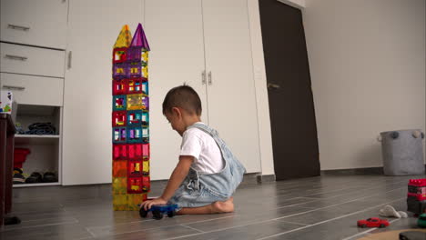 Zeitlupe-Eines-Frechen-Jungen-Lateinamerikanischen-Kleinkindes,-Das-Einen-Turm-Aus-Magnetischen-Kacheln-Mit-Spielzeugautos-Darin-Zerstört