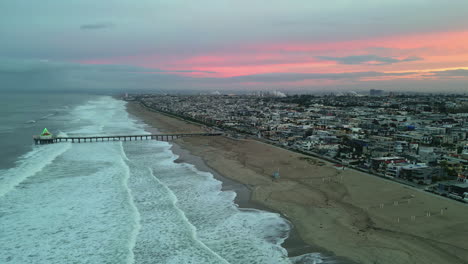 Manhattan-Beach-Ocean-Waves-with-Pier-Overlooking-an-Urban-Cityscape-with-Morning-Pink-Sunrise,-Aerial-Drone-Panning-Shot