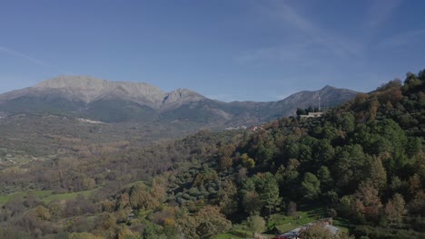 flight-in-a-valley-in-autumn-with-colorful-trees-with-pine-and-oak-forests-and-some-green-meadow-with-a-background-of-granite-mountains-and-several-towns-on-a-day-with-blue-sky-in-Avila-Spain