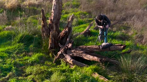 Outdoor-Photographer-With-Camera-And-Tripod-Next-To-Dead-Tree-Wood-On-Field