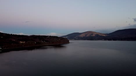 Twilight-hues-over-calm-lake-with-distant-hills-and-scattered-lights,-serene-evening-atmosphere,-aerial-view