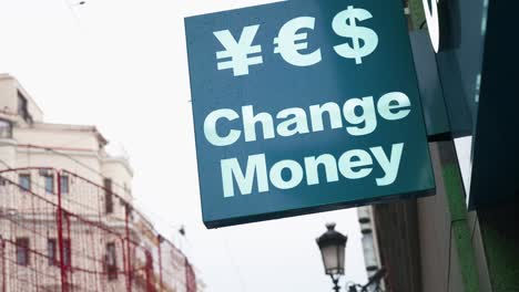 A-currency-exchange-sign-shows-the-message-'Change-Money'-with-symbols-for-currencies-like-Japanese-yen,-US-dollar,-and-euro-on-a-retail-commercial-street-in-Madrid,-Spain