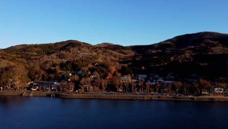 Golden-hour-view-of-a-serene-lakeside-town-with-autumnal-trees-and-hillside-homes,-clear-skies-above