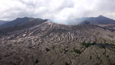 Behold-the-majestic-Bromo,-an-iconic-and-active-volcano-in-East-Java,-Indonesia,-showcasing-awe-inspiring-volcanic-activities-as-it-releases-billows-of-gray-smoke-and-ash,-aerial-4k-drone-footage