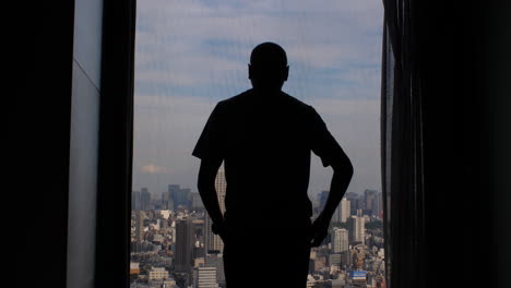 Die-Silhouette-Eines-Mannes-Geht-Auf-Ein-Großes-Fenster-In-Einem-Hochhaus-Zu-Und-öffnet-Die-Vorhänge,-Um-Den-Blick-Auf-Die-Weite-Stadtmetropole-Freizugeben